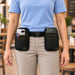 A person wearing a light blue polo shirt and khaki pants models a black waist bag set with two front POS pouches. The left pouch holds pens, and the right pouch holds a small black device.