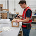 Warehouse worker wearing an AgozTech trigger scanner holster while scanning a labeled box at a packing station.