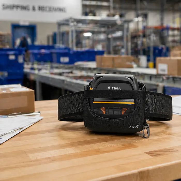 Zebra ZQ521 mobile printer waist belt carry case on a wood workbench in a logistics center, with warehouse shelving, cartons, and fulfillment equipment in the background.